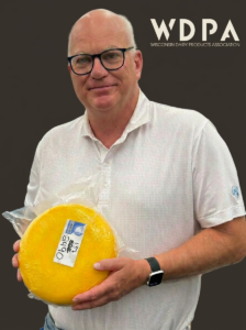 Smiling man with glasses holds a large yellow cheese wheel in plastic wrap, WDPA logo visible in the top right.