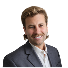 Professional headshot of a smiling man wearing a dark blazer and white shirt, facing the camera against a neutral background.