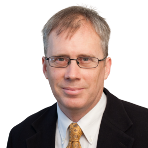 Close-up portrait of a man in a dark suit, white shirt, and patterned gold tie, wearing rectangular glasses.