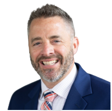 Smiling professional headshot of a man with short gray hair and a beard, wearing a dark blue suit and a pink striped tie.